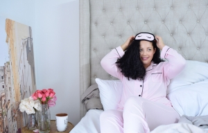 Tanesha Awasthi lounging on a Restoration Hardware bed in a pink and black pajama set with matching sleep mask from Nordstrom.
